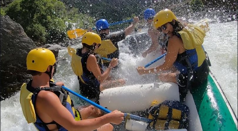 Group whitewater rafting through rocky river rapids, wearing yellow helmets and life vests as blue paddles cut through splashing water.