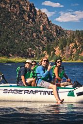 Group of four in green life jackets on an inflatable raft enjoying a sunny mountain river canyon, one person smiling and dipping a foot in the water.
