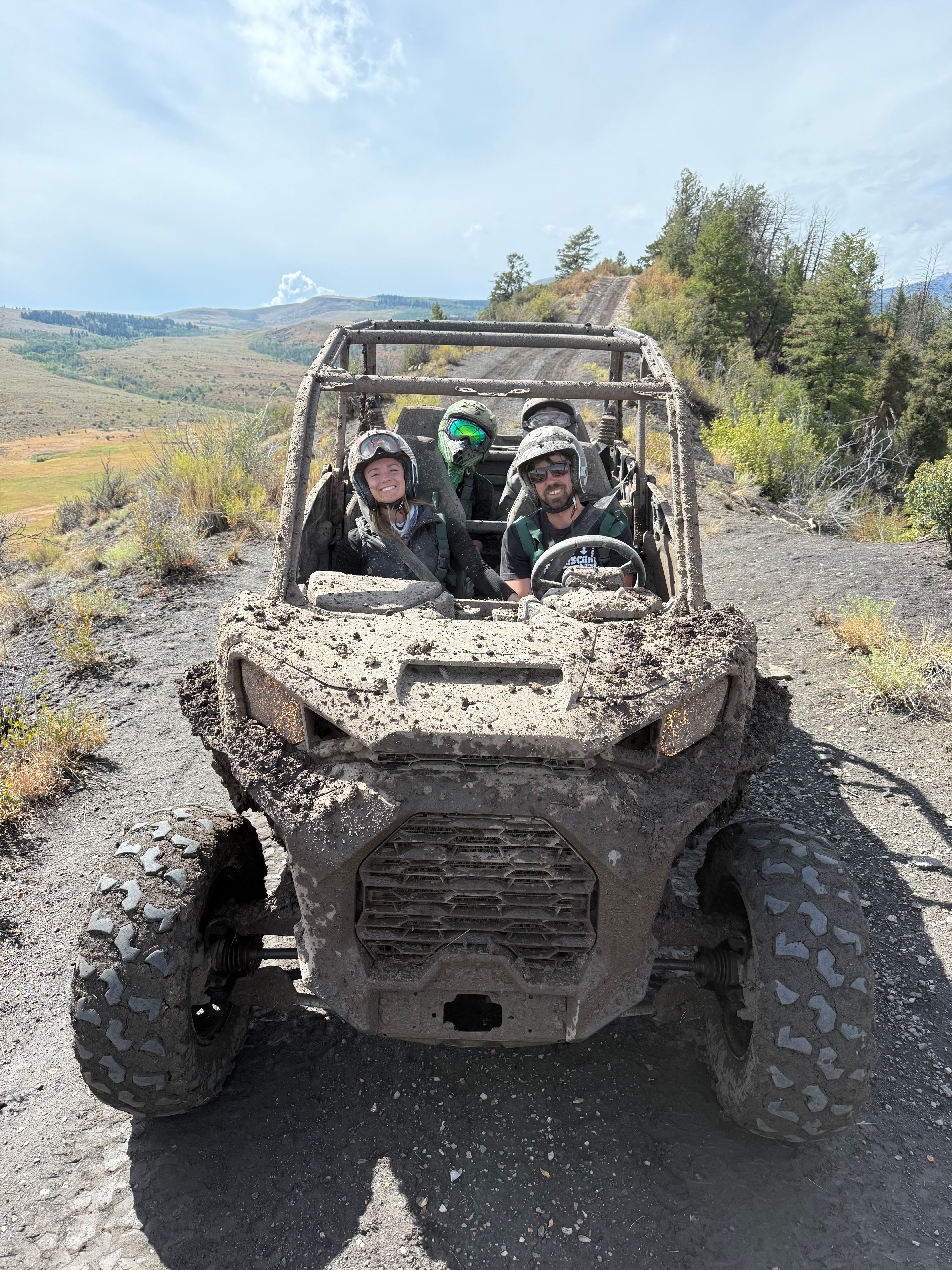 Muddy off-road UTV with three helmeted riders smiling on a mountain dirt trail, scenic rolling hills and pine trees in the background — outdoor adventure.