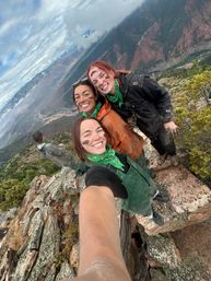 Group selfie of four hikers wearing green bandanas and muddy face paint, smiling on a rocky mountain ridge overlooking a deep canyon and cloudy sky during an outdoor adventure