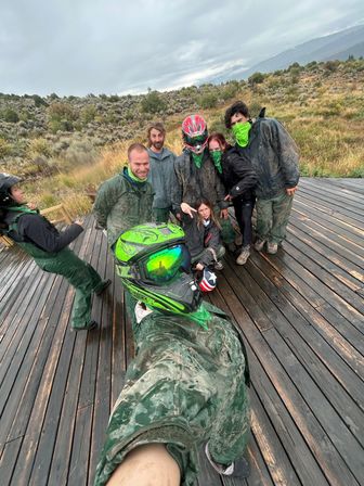 Mud-splattered group selfie of off-road riders in helmets and green bandanas on a wet wooden deck overlooking a sagebrush high‑desert landscape under a cloudy sky.