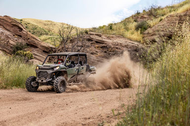 Four-seat off-road UTV racing along a dirt trail through rocky hills and dry grass, kicking up a huge dust cloud with two helmeted riders enjoying an adventurous ride.