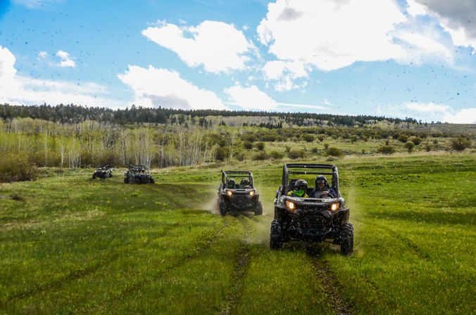 Side-by-side UTVs kicking up mud as riders drive across a green mountain meadow with a tree-lined ridge and blue sky with clouds.