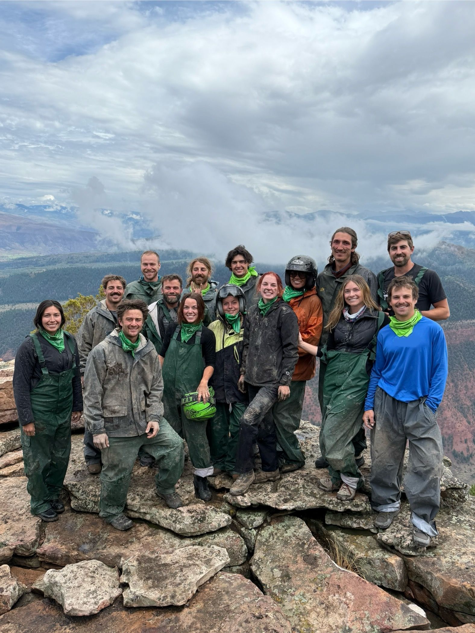 Group of 15 muddy hikers in green bandanas and outdoor gear posing on a rocky mountaintop cliff with dramatic cloudy sky and sweeping valley views.