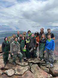 Group of 15 muddy hikers in green bandanas and outdoor gear posing on a rocky mountaintop cliff with dramatic cloudy sky and sweeping valley views.