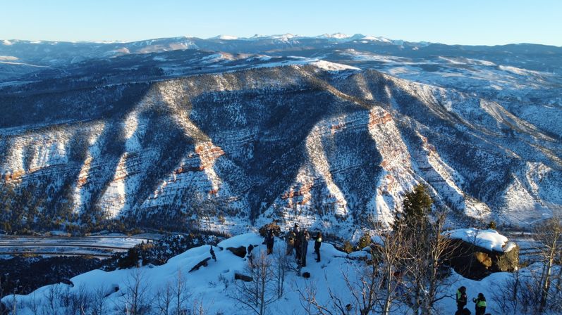 Aerial view of a snowy mountain overlook with hikers on a rocky ridge overlooking sunlit, snow-covered canyons and distant peaks at winter sunrise.