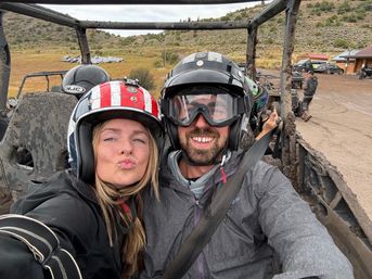 Two helmeted riders smiling for a selfie in a muddy off-road UTV on a grassy hillside dirt trail