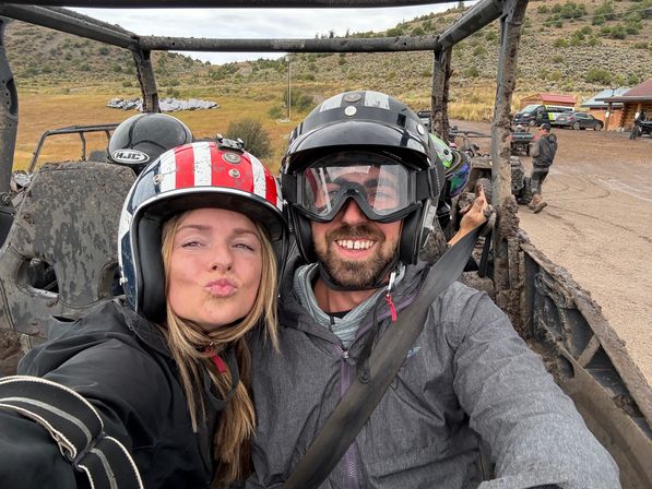 Two helmeted riders smiling for a selfie in a muddy off-road UTV on a grassy hillside dirt trail
