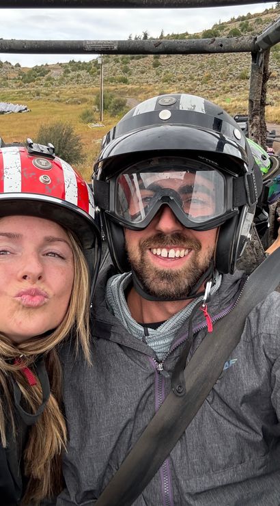 Two helmeted riders smiling for a selfie in a muddy off-road UTV on a grassy hillside dirt trail