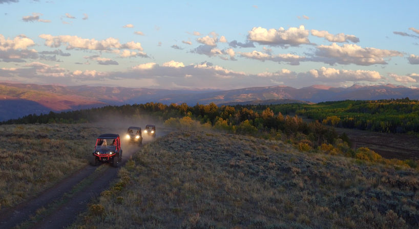 Four off-road UTVs kicking up dust on a dirt trail along a grassy ridge with golden aspens and layered mountain ranges under a pastel sunset sky.