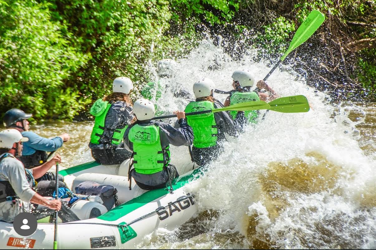 Group of rafters in green life jackets and white helmets paddling through a splashy whitewater rapid on a tree-lined river