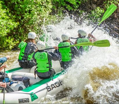 Group of rafters in green life jackets and white helmets paddling through a splashy whitewater rapid on a tree-lined river