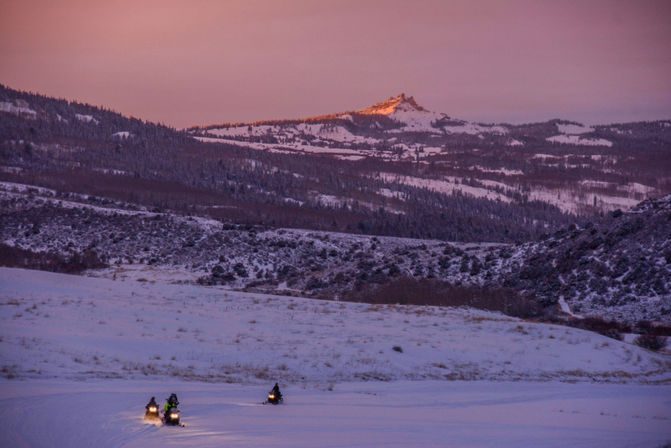 Three snowmobilers with headlights crossing a snow-covered valley at dusk, pink alpenglow sky and a sunlit mountain peak in the distance.