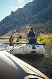 Paddlers on an inflatable raft drifting down a mountain river with pine-covered cliffs, sunny sky, and a relaxed summer rafting adventure