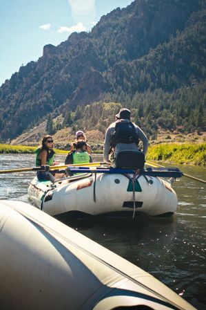 Paddlers on an inflatable raft drifting down a mountain river with pine-covered cliffs, sunny sky, and a relaxed summer rafting adventure