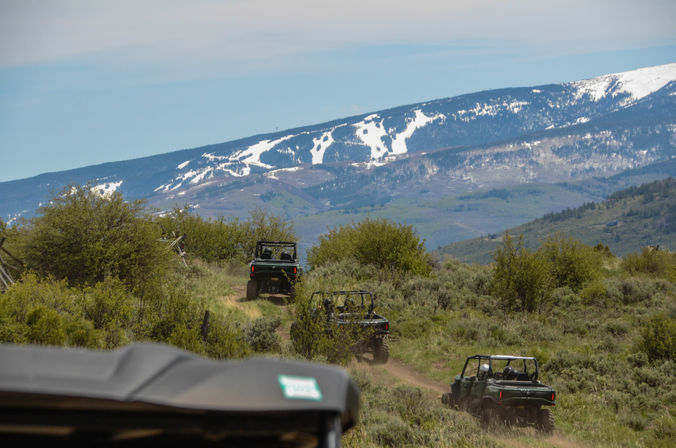 Several off-road UTVs riding single-file on a dusty trail through sagebrush and green foothills with snow-dusted mountains and a blue sky in the background — scenic outdoor adventure.