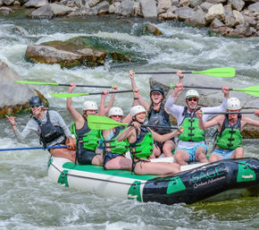 Group of rafters in green life jackets and helmets raising bright green paddles while navigating rocky whitewater rapids on a mountain river