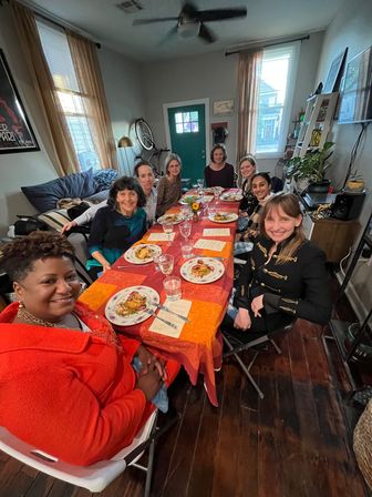 Group of friends enjoying a cozy dinner party around a long orange-red tablecloth in a sunlit city apartment, plated meals and wine glasses on the table, hardwood floors, potted plants and a green entry door visible.