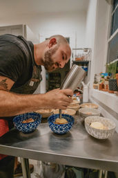 Person in apron plating desserts in a cozy home kitchen, using a metal culinary dispenser to pipe cream into blue patterned bowls on a stainless-steel prep table.