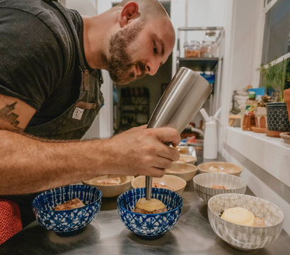 Person in apron plating desserts in a cozy home kitchen, using a metal culinary dispenser to pipe cream into blue patterned bowls on a stainless-steel prep table.