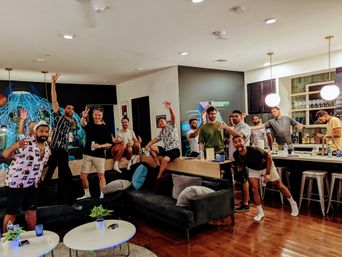 Group of young men enjoying a lively house party in a modern open-plan apartment living room and kitchen, posing with drinks on a sectional sofa and around a kitchen island under pendant lights and a colorful mural