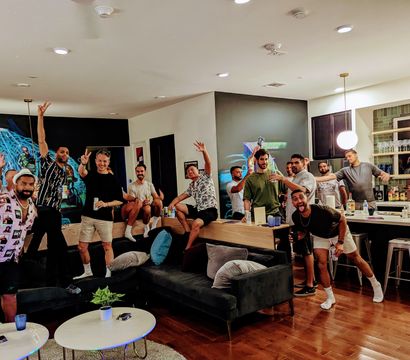 Group of young men enjoying a lively house party in a modern open-plan apartment living room and kitchen, posing with drinks on a sectional sofa and around a kitchen island under pendant lights and a colorful mural