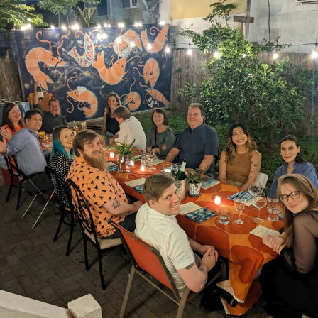 Outdoor backyard dinner party at night with a long orange-tablecloth table, string lights and candles, wine glasses, and a smiling group seated beneath a colorful shrimp mural.