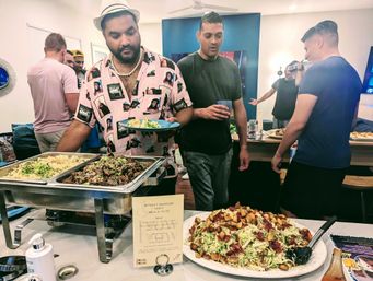Casual indoor party buffet with friends grabbing plates of rice, sesame-topped beef and a large crouton-topped salad from catering trays.