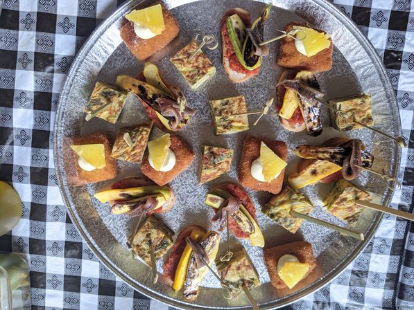 Silver platter of assorted bite-size tapas: golden croquettes topped with cream and pineapple, herb omelet squares, and skewers of grilled vegetables and anchovy-style fillets, on a black-and-white checkered tablecloth.