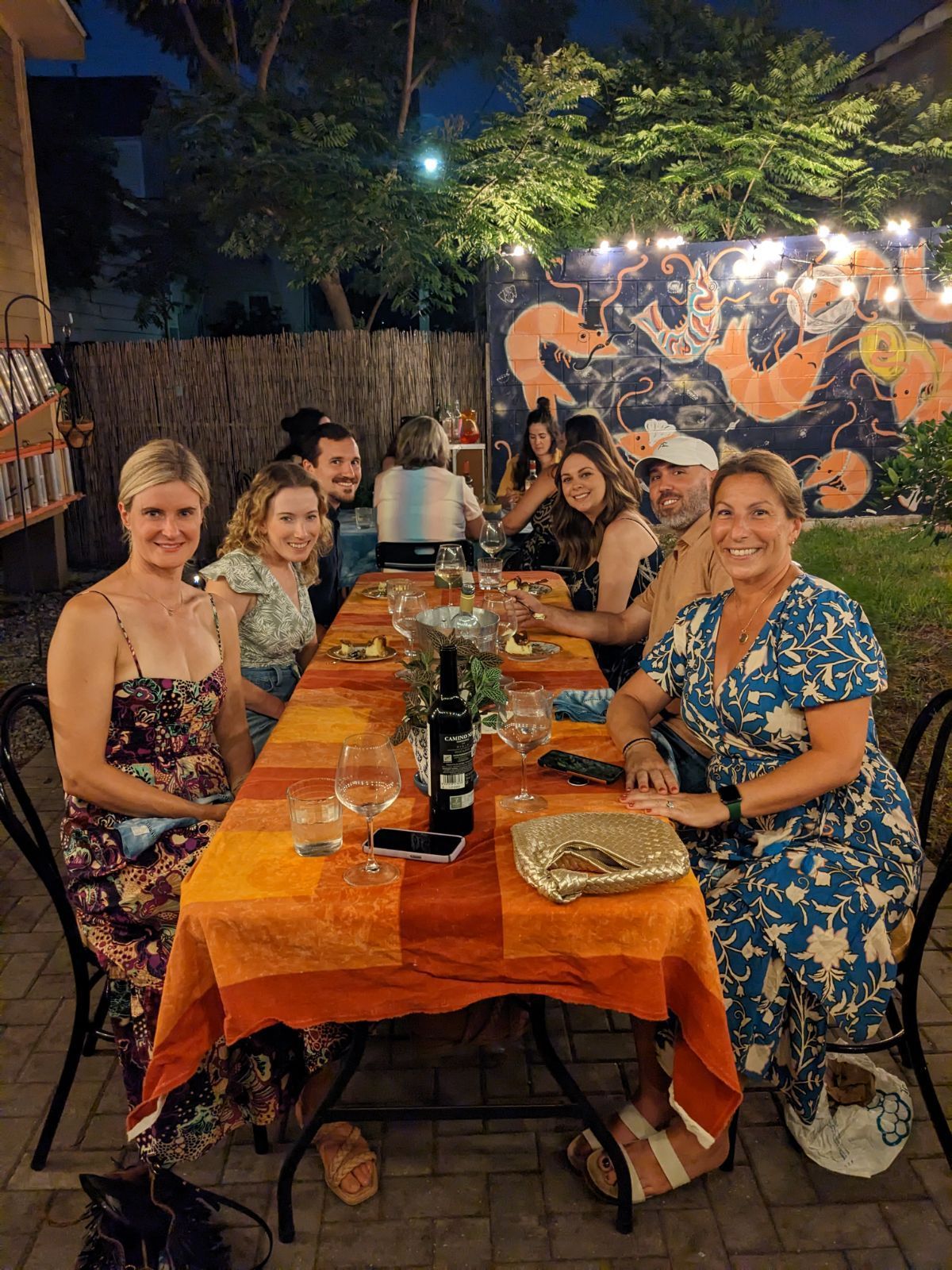 Smiling friends enjoying a casual outdoor backyard patio dinner at night — long orange tablecloth with wine glasses, string lights and a colorful sea‑creature mural on the fence.
