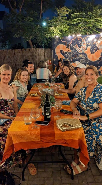 Smiling friends enjoying a casual outdoor backyard patio dinner at night — long orange tablecloth with wine glasses, string lights and a colorful sea‑creature mural on the fence.