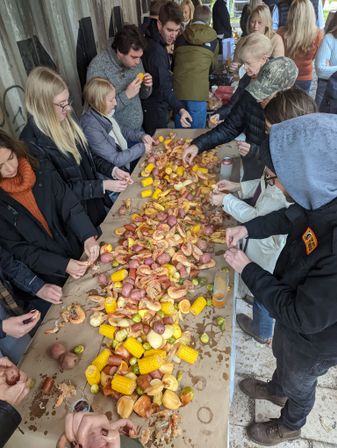 Communal outdoor seafood boil on a long paper-covered table: piled shrimp, corn on the cob, red potatoes, sausage and veggies with a crowd in jackets gathered around digging in.