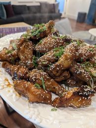 Steaming sesame-glazed chicken wings sprinkled with white sesame seeds and sliced scallions on a decorative white plate in a cozy living-room setting.