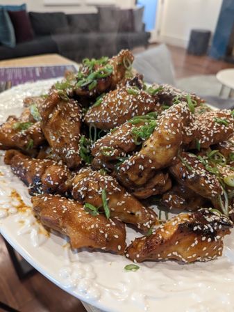 Steaming sesame-glazed chicken wings sprinkled with white sesame seeds and sliced scallions on a decorative white plate in a cozy living-room setting.