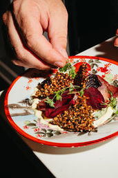 Hand garnishing a gourmet beet and seed-crusted croquette salad with microgreens on a decorative red-rimmed plate, plated restaurant appetizer.