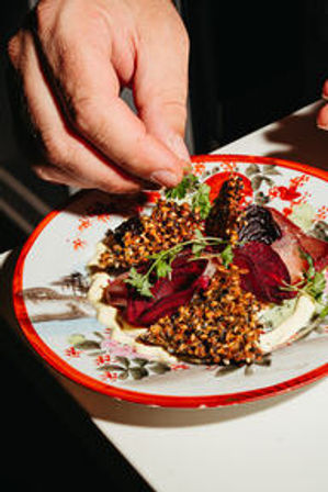 Hand garnishing a gourmet beet and seed-crusted croquette salad with microgreens on a decorative red-rimmed plate, plated restaurant appetizer.