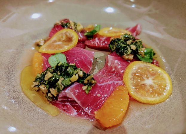 Vibrant tuna crudo with kumquat and mandarin citrus, chopped herb-and-nut gremolata and microgreens on a beige fine-dining plate.