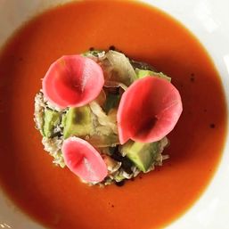 Top-down view of a gourmet roasted tomato soup in a white bowl, garnished with a central mound of diced avocado and shredded seafood, topped with three bright pink pickled radish petals.