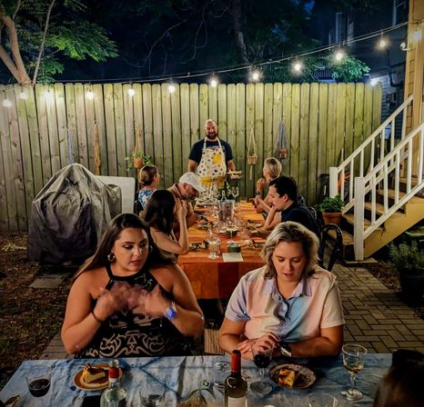 Cozy backyard patio dinner at night with friends around a long table, wine and dessert under string lights by a wooden fence and porch steps.