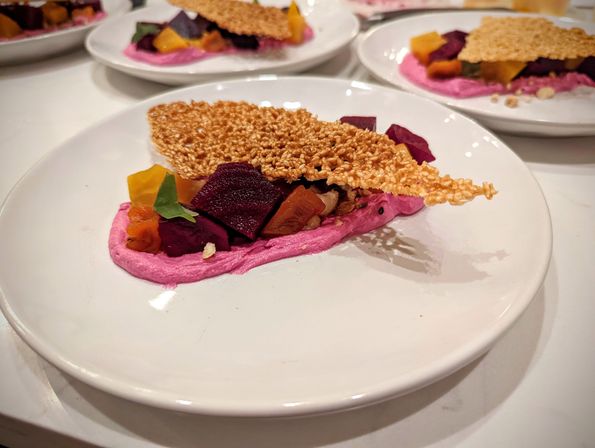 Fine-dining plated appetizer: vibrant pink beet purée with roasted red and golden beet cubes, a crunchy sesame tuile and microgreens on a white plate.