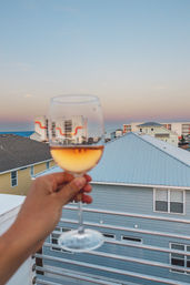 Hand holding a glass of rosé on a beachfront balcony at sunset, pastel beach houses and low-rise oceanfront condos under a gradient pink-blue sky.