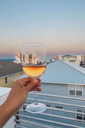 Hand holding a glass of rosé on a beachfront balcony at sunset, pastel beach houses and low-rise oceanfront condos under a gradient pink-blue sky.