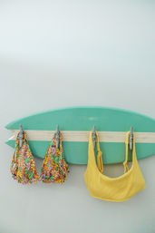 Mint-green wall-mounted surfboard used as a rack with metal hooks holding a floral bikini top and a yellow ribbed bikini top — beach house summer decor.