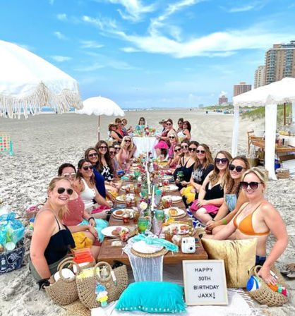 Cheerful seaside beach picnic: a large group of women seated at a long low table on a sandy oceanfront beach under white umbrellas, colorful plates and drinks, a 30th birthday sign, high-rise shoreline and blue sky