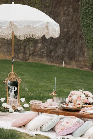 Boho-chic garden picnic on a green lawn with a fringed white umbrella, low wooden table with plates and charcuterie, pink-and-white floral centerpiece, and plush pillows and blankets.
