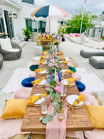 Boho-chic low wooden table set for a summer waterfront brunch on a coastal deck — colorful round cushions, mustard napkins, glassware, floral centerpieces and parasol.