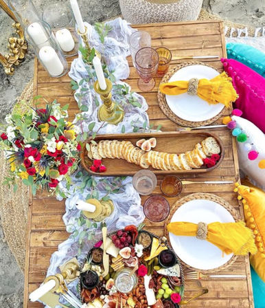 Boho beach picnic low wooden table on sand with charcuterie platter of crackers, cheeses, figs and grapes, woven place settings with yellow napkins, brass candlesticks, candles, colorful pom-pom cushions and a floral arrangement.