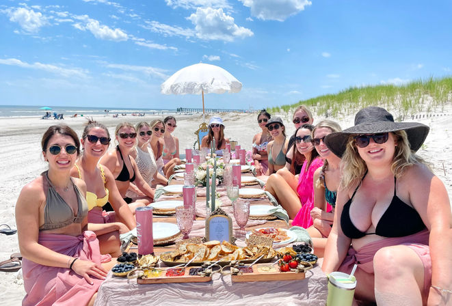 Group of friends enjoying a sunny coastal beach brunch: a long low table on the sand with pink linens, platters of bread, fruit and drinks, a white parasol, sand dunes and a pier under a blue sky.