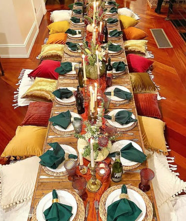 Long low wooden table set for a cozy bohemian autumn dinner with woven chargers, green napkins, brass candlesticks, wine bottles and colorful velvet floor pillows on polished hardwood