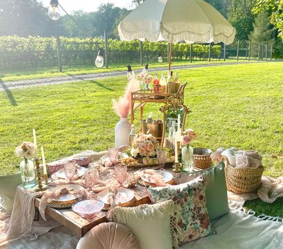 Boho-chic outdoor picnic in a sunlit vineyard: low wooden table on blankets with pastel plates and crystal glasses, floral cushions, candles, wicker basket, fringed umbrella and string lights.